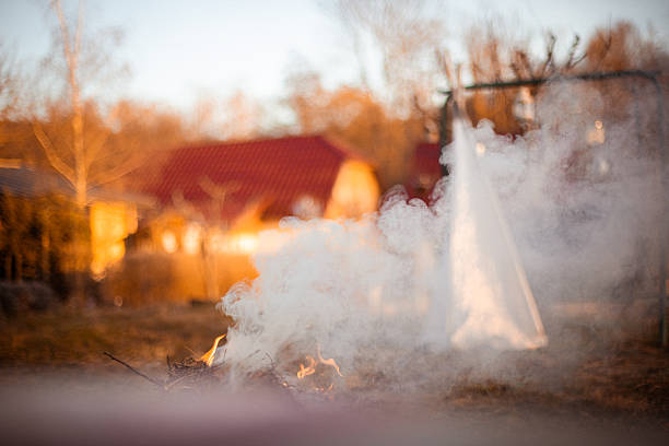 Atmospheric outdoor scene with rising smoke and small fire in a rural backyard during golden hour. Warm sunset light, soft focus, and drifting smoke create a moody, cinematic feeling. Concept of transition, mystery, rural life, seasonal change, and environmental atmosphere.