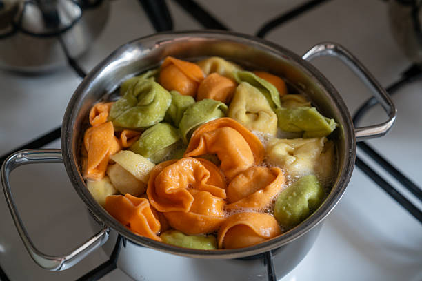 Close-up shot of green, orange, and yellow tortellini pasta boiling in a stainless steel pot on a white gas stove. Concept of home cooking, Italian cuisine, and preparing a healthy meal.