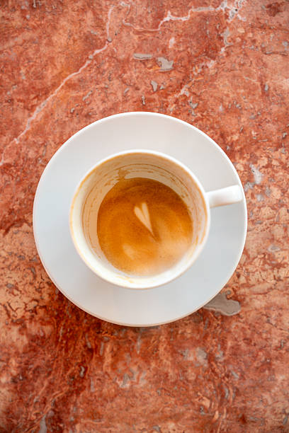 A high-angle shot of an almost empty white ceramic coffee cup and saucer on a textured pink marble surface. The bottom of the cup shows the remains of a latte art heart, symbolizing the end of a pleasant coffee break. Minimalist and aesthetic composition for lifestyle and food content.