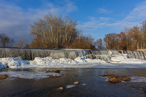 Breathtaking shots of a frozen dam on the Jihlava River. Massive ice formations, icicles, and frozen waterfalls covering the weir structure. The contrast between flowing water and thick ice creates a dramatic winter landscape under a sunset sky