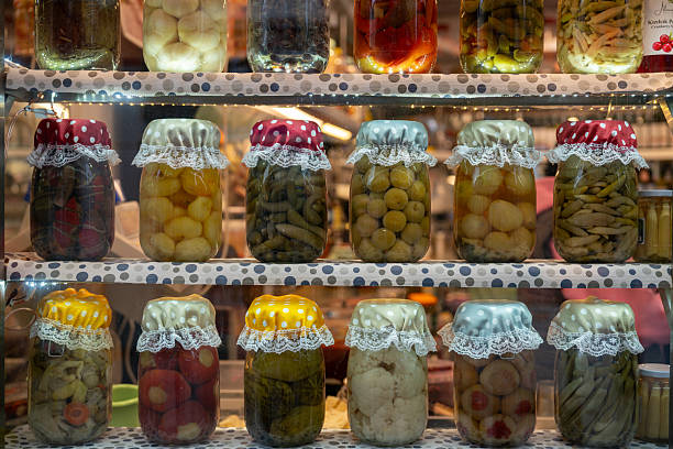 A front-view shot of various homemade pickled vegetables in glass jars, neatly arranged on shelves. Each jar is decorated with colorful polka-dot fabric and lace-trimmed lids. Includes pickled peppers, cucumbers, cauliflower, and tomatoes. Perfect for traditional food, fermentation, and autumn harvest themes