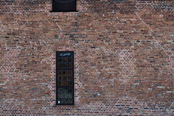 Close-up view of a red brick wall featuring a narrow vertical window, showcasing minimalist urban architecture and textured masonry. Ideal for concepts of simplicity, isolation, construction, building exterior, renovation, history, or industrial design. Horizontal composition with copy space.