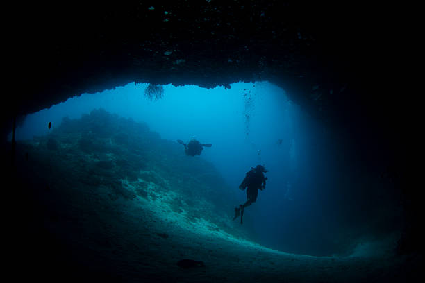 Two divers swim in a dark underwater cave while exploring the ocean floor. The scene shows clear blue water and rock formations. Sunlight filters through the cave entrance.