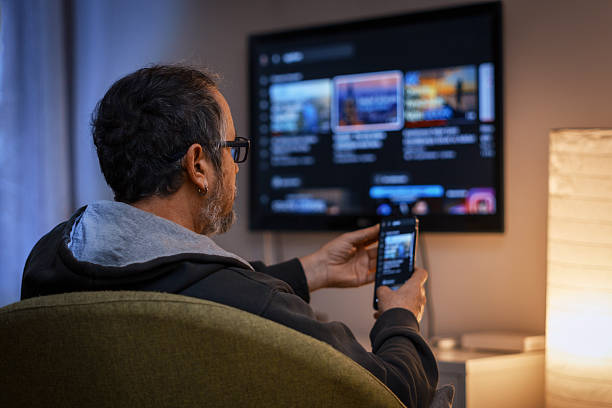 Mature man relaxing at home watching streaming content on smart TV using smartphone