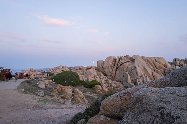Capo Testa, Italy - September 28, 2023: Several people are walking along a dirt path beside large granite rock formations and low vegetation at Capo Testa during dusk, with the moon visible in the sky.