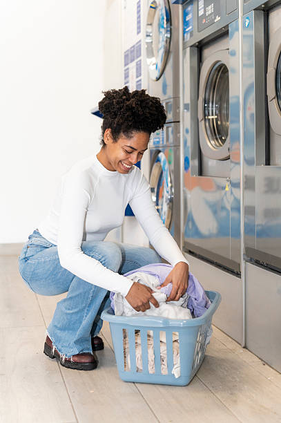 Smiling woman doing routine laundry chores, putting clothes into a washing machine at a modern self-service laundromat