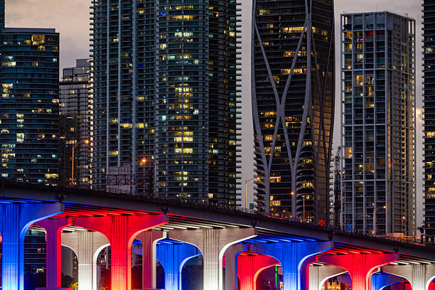 This is a photograph of the downtown Miami skyline at dusk with the MacArthur Causeway bridge illuminated in red, white and blue colors.