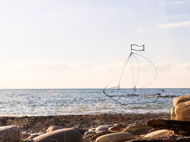 Blue wireframe sketch of a sailboat with flag on calm waters, foregrounded by pebble beach and soft evening sky.