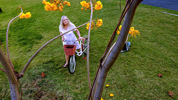 Mature woman pushes bicycle yellow blossoms near canal, Bangkok