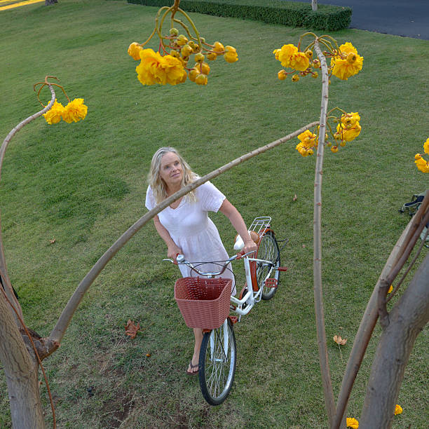 Mature woman pushes bicycle yellow blossoms near canal, Bangkok