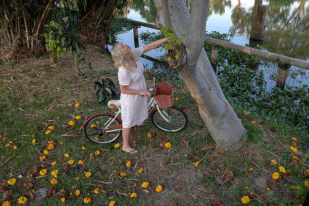 Mature woman pushes bicycle yellow blossoms near canal, Bangkok