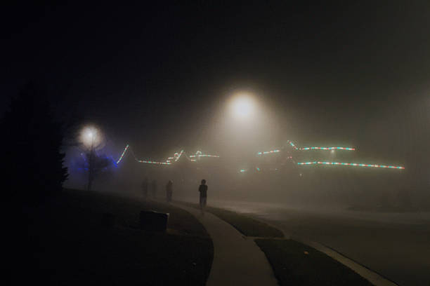 Silhouetted figures are visible in the light of a streetlight on a foggy night. Christmas lights are visible in the background
