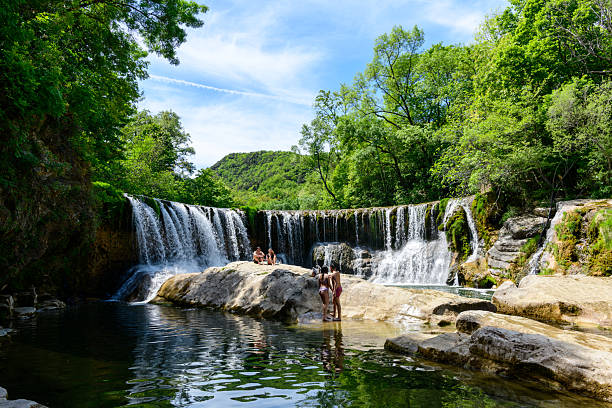 Saint Laurent le Minier, France - May 6, 2023: Several people are visible standing and sitting on rocks in front of the Cascade de la Vis waterfall, surrounded by green trees and natural vegetation under a blue sky.