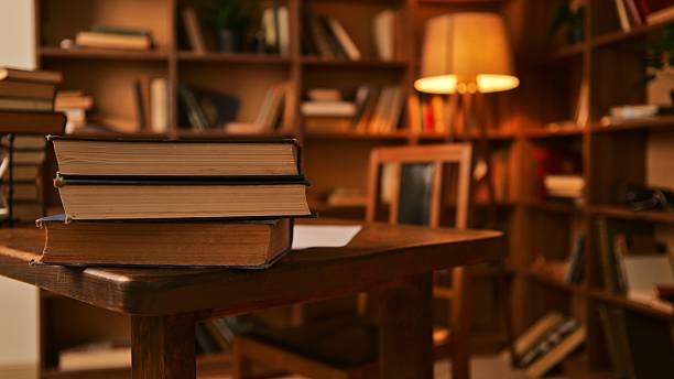 A warm, inviting scene unfolds in a library, featuring a stack of old books on a table, with bookshelves and a glowing lamp in the background