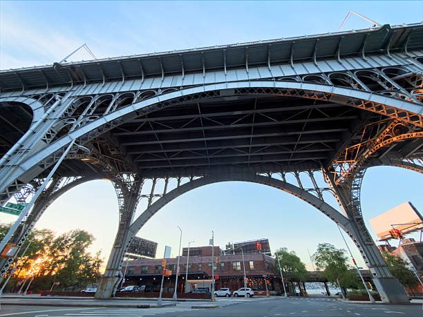 Low perspective shot looking up at the steel arches and beams of an elevated highway as it spans across a city street, with apartment buildings, trees and evening sky visible in the background for an urban infrastructure scene.​The dramatic underside view, converging lines and mix of traffic routes and neighborhood architecture make this image ideal for concepts related to transportation, city planning, civil engineering and gritty New York street life.