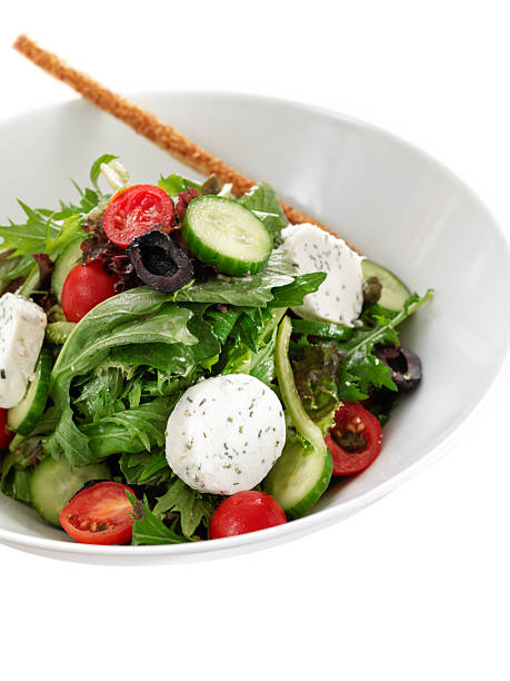 Overhead view of a fresh mixed green salad with goat cheese rounds, cherry tomatoes and breadstick served in a white ceramic bowl on a clean white background, styled as an isolated food image with generous copy space for text or logo placement.​​The vibrant green leaves, red tomatoes and creamy cheese create a colorful, appetizing composition ideal for healthy eating, vegetarian cuisine, restaurant menu design and modern food photography concepts.