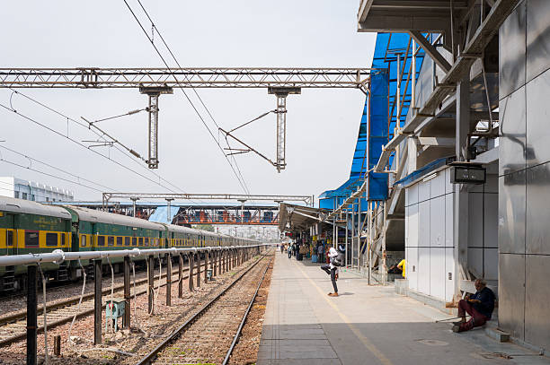Delhi, India - March 10, 2024: Passengers wait on the platform at Hazrat Nizamuddin railway station, with a green and yellow long-distance train on the tracks, overhead electric wires, and blue pedestrian bridges visible.
