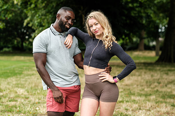 Young interracial couple is standing together in a park, smiling at the camera with the woman's arm around the man's shoulder and the man's arm around the woman's waist