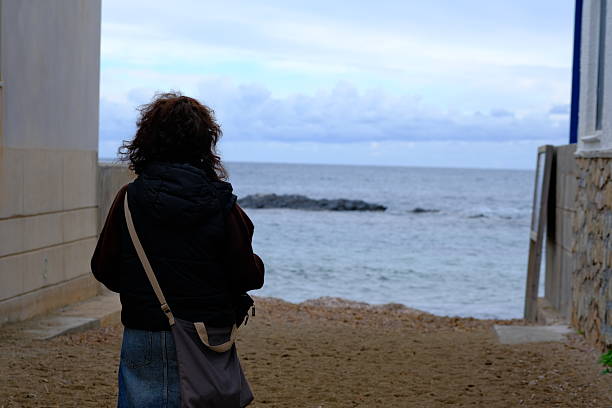 A woman walks towards the beach from an alley in a coastal town on a cold winter afternoon, with calm water.