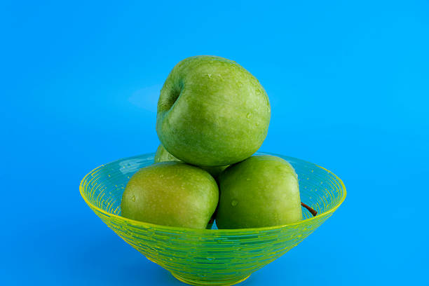 A glass bowl filled with fresh green apples, arranged in a pyramid shape, set against a vibrant blue background. A simple and fresh fruit composition