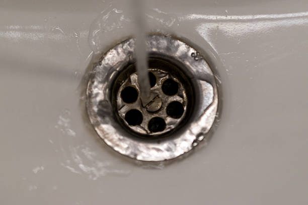 Close-up view of clean running water flowing into a metal sink drain in a bathroom. The image represents household hygiene, daily routine, plumbing, and domestic water usage with a minimal and neutral interior background.