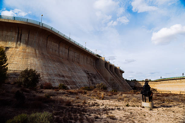 Impressive concrete dam spillway curves like a cliff, with a dark metal valve sculpture in the foreground. Dry shrubbery and a clear blue sky create a stark, engineering-inspired landscape.