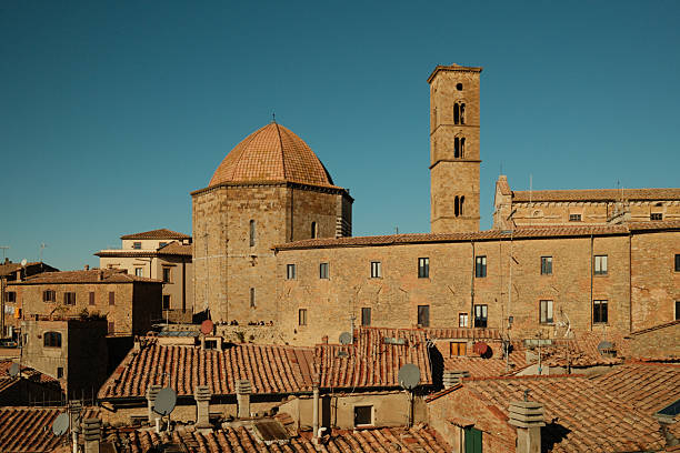 Volterra cityscape presenting the historic Baptistery of San Giovanni and a bell tower under a clear blue sky, showing traditional terracotta roofs