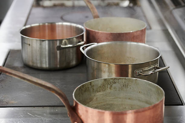 Close-up view of copper and stainless steel saucepans arranged on a professional kitchen stove. The worn surfaces and patina highlight frequent use and craftsmanship, creating an authentic culinary still life. Selective focus and neutral tones emphasize texture, depth, and a working kitchen environment.