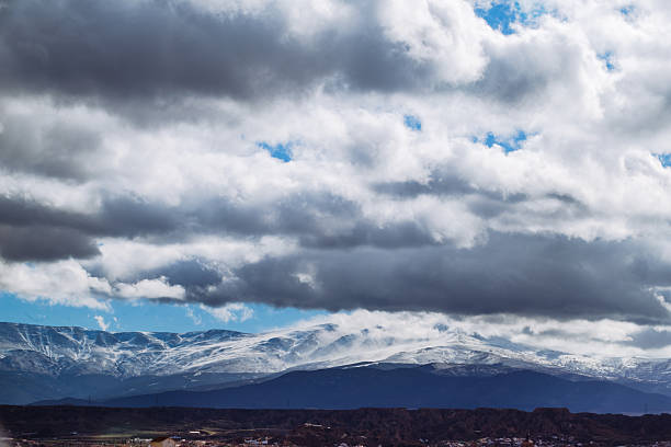 A breath-taking scene featuring dark blue mountain hills adorned with snowy tops, set against a backdrop of captivating grey clouds.
