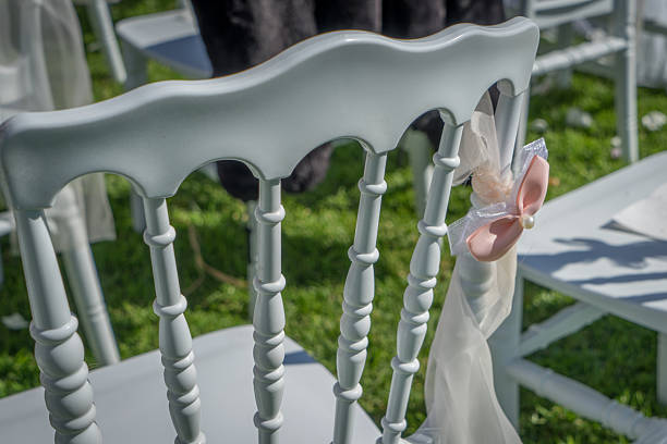 White wedding chair adorned with a delicate pink bow and pearl detail set on lush green grass, perfect for elegant outdoor ceremonies, receptions, and romantic spring or summer events