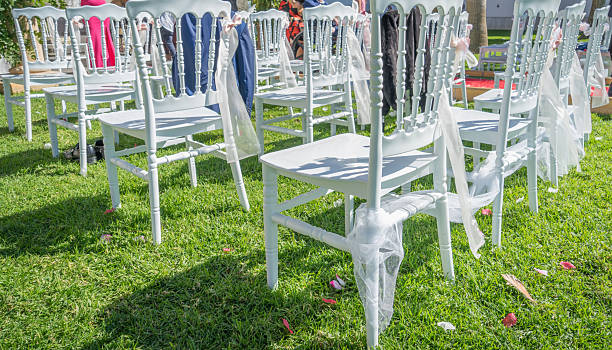 White chiavari chairs decorated with sheer fabric and scattered rose petals are arranged on green grass, preparing for an elegant outdoor wedding or special event