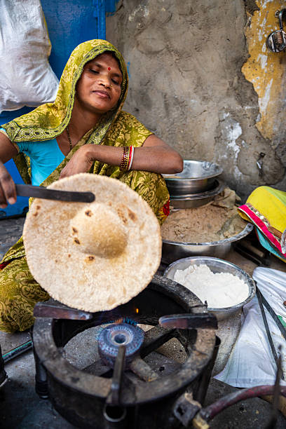 A woman in a green patterned sari and headscarf sits by a small gas burner, using a metal spatula to cook and puff a large flatbread. She wears red bangles and a red bindi, and several metal bowls of flour and dough are arranged nearby. The scene is intimate and candid, with warm colors, a worn textured wall behind her, and the focus on the hands-on process of traditional bread-making.