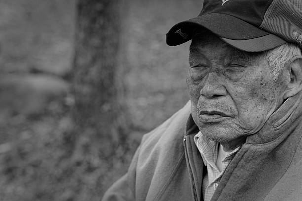 Taoyuan, Taiwan - March 10, 2024: Close-up black-and-white portrait of a weathered elderly man with closed eyes, wearing a cap and padded jacket, leaning against a tree trunk in quiet repose, evoking everyday moments of rest in Taiwan.
