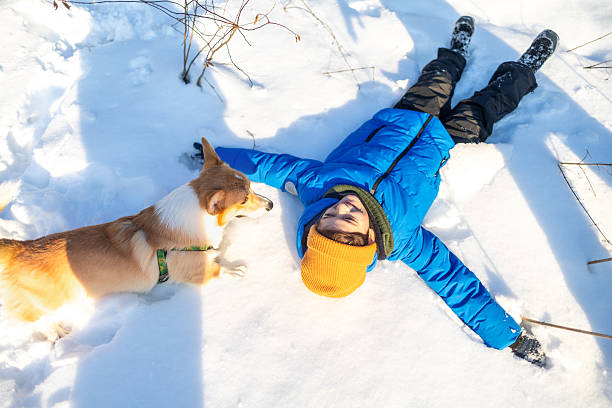 Young boy lying in the snow with his arms spread out while playing with a corgi dog during a sunny winter day. Child enjoying outdoor winter activities with a pet in a snowy forest or park, creating snow angels and bonding with a dog. Concept of childhood, friendship, winter fun, family time, pets, nature, and outdoor lifestyle.