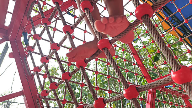 Child climbing a rope net playground structure outdoors, close-up of bare feet and legs, showing active play, balance, and motor skill development in a colorful playground environment
