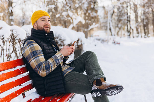 Young adult man wearing a yellow beanie and winter clothing, sitting on a park bench using a mobile phone, surrounded by snow