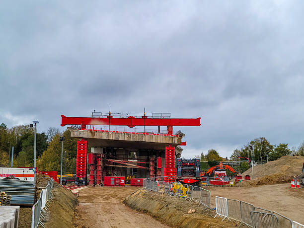 A massive concrete bridge deck is elevated by multiple red hydraulic jacks on a construction site. The structure features steel beams and railings, supported on uneven dirt ground with scattered rocks and gravel. In the background, bare earth mounds and construction debris are visible, along with a metal light pole. Surrounding the site are trees with autumn leaves in shades of yellow and orange under a cloudy gray sky. The scene depicts an ongoing infrastructure project in a rural outdoor setting during fall daytime. No people are present in the image.
