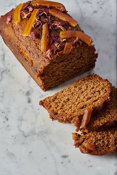 Overhead views and close-up shots of a gluten free gingerbread loaf topped with candied citrus peel and dried flower petals. The cake is sliced to reveal a dense and moist crumb, photographed on a light marble surface with natural light. Minimal styling emphasizes texture, baking details, and artisanal dessert concepts.