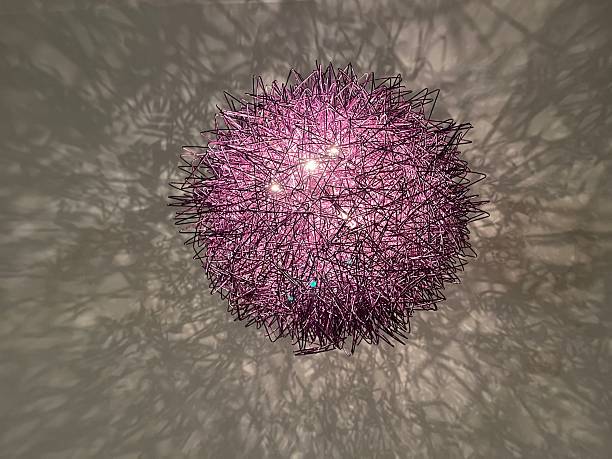 A modern purple wire sphere light fixture hangs, casting intricate, abstract shadows across a light colored, textured wall