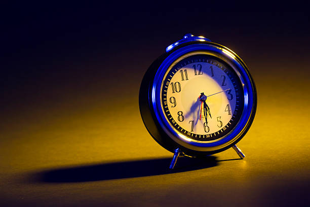 Studio still life of a classic analog alarm clock illuminated by dramatic directional lighting, casting a long shadow across a vibrant yellow surface against a dark, minimal background. The clock face glows softly while cool blue highlights contrast with warm tones, creating a cinematic and timeless mood.