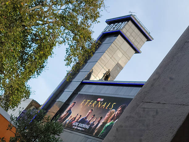 London.UK: Prominent billboard displaying the Marvel Studios film Eternals on the exterior of the Odeon Piccadilly. The poster showcases the ensemble cast including actors in superhero attire against a cosmic backdrop with the movie title in bold yellow lettering. The structure features a metallic silver and blue façade with the Odeon logo illuminated on the rooftop tower. Surrounding the building are green leafy trees and a clear blue sky, indicating an outdoor daytime setting in late summer or early fall. No people are visible in the scene. The image captures the promotional display for the 2021 superhero film.