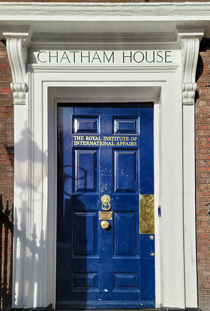 London, UK: A circular blue plaque mounted on the red brick facade of Chatham House, the Royal Institute of International Affairs, commemorates William Pitt the Younger, British Prime Minister from 1759 to 1806, who resided in this building. The plaque inscription reads 'William Pitt the Younger 1759-1806 Prime Minister lived here.' The Georgian-style architecture includes white stone cornices, a dark blue front door with brass knocker, white sash windows with blinds, black wrought-iron railings, and potted yellow flowers on the steps. The scene is set outdoors in an urban environment during daytime with natural light illuminating the structure. No people are visible in the image.