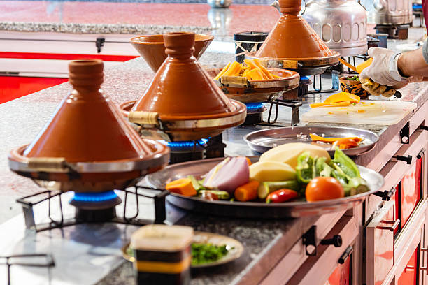 A busy Moroccan kitchen scene with clay tagine pots on stoves, a chef slicing peppers and vegetables on the counter, vibrant colors and traditional cookware creating lively culinary atmosphere.