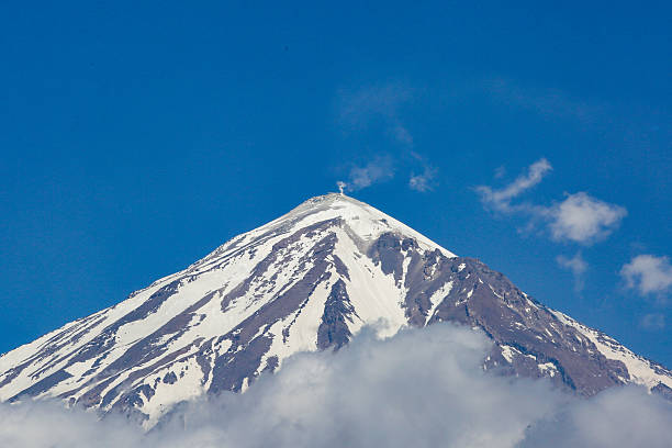 Sulfurous gas rising from the summit of Mount Damavand, the highest volcano in the Middle East, indicating ongoing geothermal activity. Snow-covered slopes contrast with fumarolic emissions under a deep blue sky, highlighting the dormant yet active nature of this iconic stratovolcano in Iran. A powerful representation of geology, volcanic activity, natural forces, and Earth's dynamic processes.