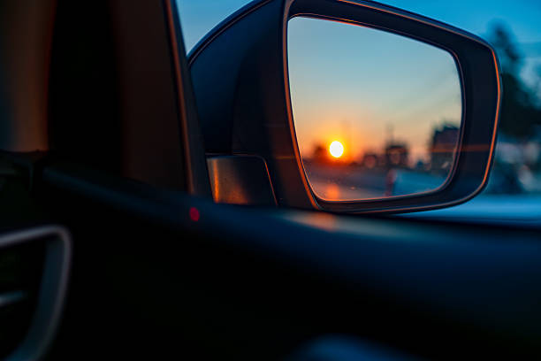 A vibrant golden sunset is captured in the reflection of a car's side-view mirror, showcasing the beauty of a golden hour commute. The background features blurred vehicles in traffic, emphasizing a journey or a road trip during the transition from day to night.