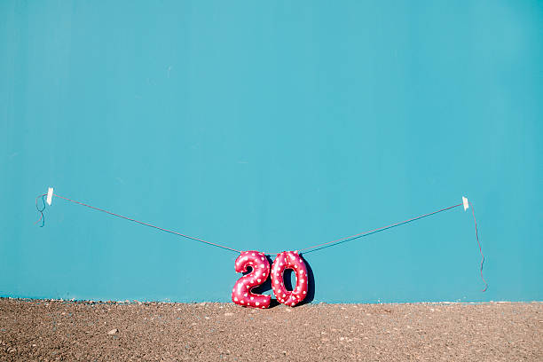 the number 20, created with pink heart patterned foil balloons, hangs from a thin red string against a wide blue wall under clear daylight