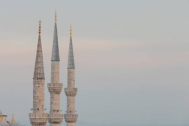Minimalist composition focusing on three slender mosque minarets rising into a pale sky. The clean background emphasizes vertical lines, architectural form, and the symbolic presence of Islamic religious structures.