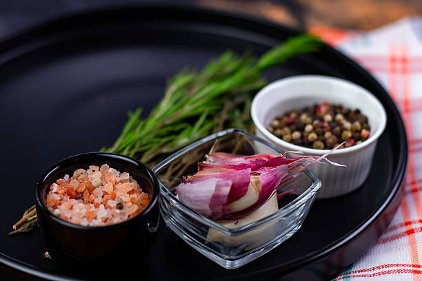 Fresh herbs and spices are displayed on a black plate. There are bowls of pink salt, various peppercorns, and sliced onion. This arrangement is for meal preparation in a kitchen.