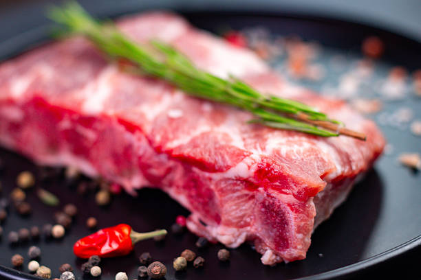 Fresh cut of meat rests on a black plate surrounded by spices and herbs. The scene highlights a raw ingredient for grilling or cooking in a kitchen setting.