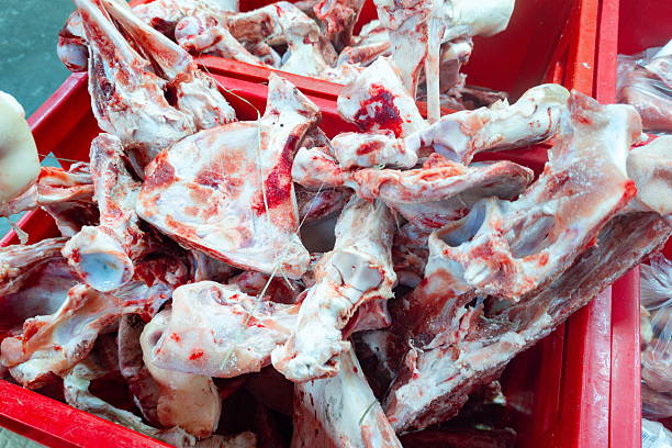 Meat bones are piled in a red container at a market. Vendors prepare their stalls for customers as the day begins. The scene shows the freshness of the products available for sale.